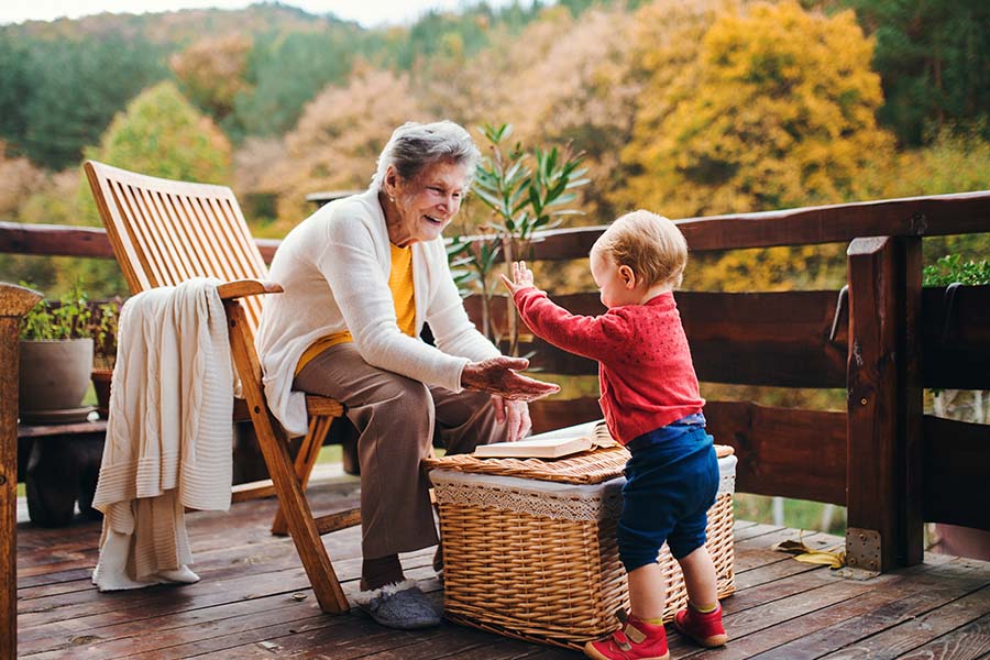 About Our Agency - Portrait of a Cheerful Grandmother Sitting on a Chair on the Back Deck While Having Fun Playing with Her Young Grandson on a Sunny Fall Day